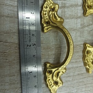 Op de afbeelding: Goudkleurige sierlijke metalen ladegreep met een decoratief ontwerp. De greep is ongeveer 6,5 cm lang en heeft twee gaten voor bevestiging.