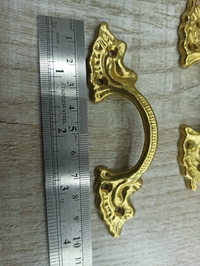 Op de afbeelding: Goudkleurige sierlijke metalen ladegreep met een decoratief ontwerp. De greep is ongeveer 6,5 cm lang en heeft twee gaten voor bevestiging.