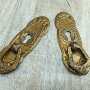May include: Two antique gold-toned metal door pulls with keyhole cutouts and decorative edges. Each pull has a loop handle.