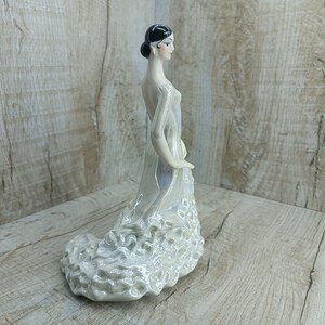 K&ouml;nnte beinhalten: Eine wei&szlig;e Porzellanfigur einer Frau in einem flie&szlig;enden Kleid. Das Kleid hat einen ger&uuml;schten Rock und ist mit einem Perlmuttglasur verziert. Die Frau hat schwarze Haare und tr&auml;gt einen kleinen Ohrring.