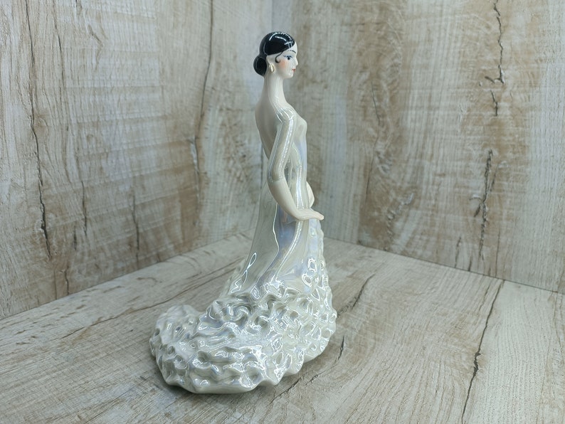 K&ouml;nnte beinhalten: Eine wei&szlig;e Porzellanfigur einer Frau in einem flie&szlig;enden Kleid. Das Kleid hat einen ger&uuml;schten Rock und ist mit einem Perlmuttglasur verziert. Die Frau hat schwarze Haare und tr&auml;gt einen kleinen Ohrring.