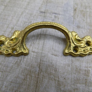 Pode incluir: Puxador de gaveta de metal dourado com um design decorativo e ornamentado. O puxador tem uma forma curva e duas extremidades decorativas.