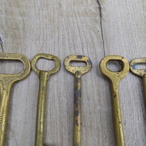 May include: Five antique brass keys with different shapes and sizes. The keys are arranged in a row on a light brown wooden surface.