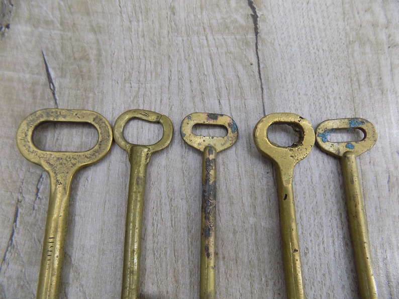 May include: Five antique brass keys with different shapes and sizes. The keys are arranged in a row on a light brown wooden surface.