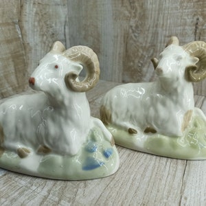 May include: Two white porcelain figurines of rams with brown horns, lying down on green bases with brown and blue accents.