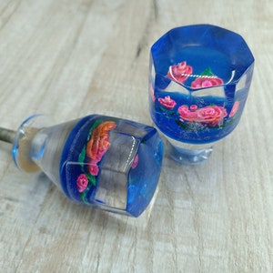 Puede incluir: Dos pomos de armario de vidrio azul con flores rosas y naranjas en su interior. Los pomos tienen forma octogonal y tienen un tornillo en el lateral.