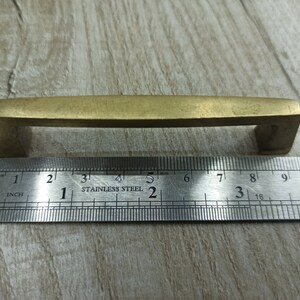 Op de afbeelding: Een messing kastgreep van 7,6 cm lang. De greep heeft een rechthoekige vorm met afgeronde hoeken en een licht gebogen bovenkant.