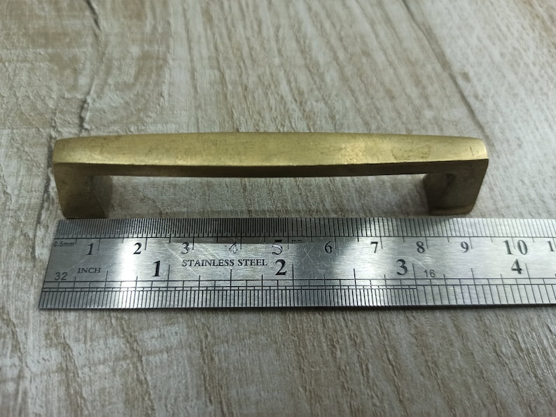 Op de afbeelding: Een messing kastgreep van 7,6 cm lang. De greep heeft een rechthoekige vorm met afgeronde hoeken en een licht gebogen bovenkant.