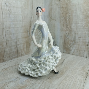 K&ouml;nnte beinhalten: Eine Porzellanfigur einer Frau in einem wei&szlig;en und silbernen, gerafften Kleid, die einen F&auml;cher h&auml;lt. Sie hat dunkles Haar und eine rosa Blume im Haar.