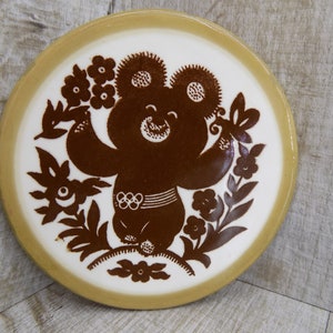 Puede incluir: Un plato de cerámica marrón y blanco con una silueta marrón de un oso que lleva un cinturón de anillos olímpicos y rodeado de diseños florales.