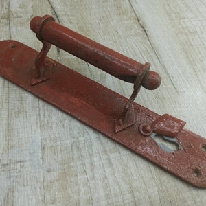 Puede incluir: Una manija de puerta de metal rojo oxidado con una placa rectangular y una manija de palanca. La manija tiene una cerradura y dos pequeños agujeros en la placa.