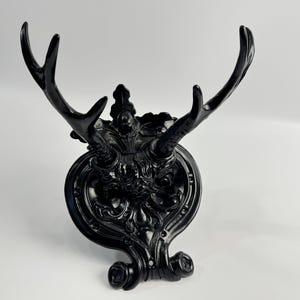 May include: A black decorative wall mount featuring a stylized antler design. The wall mount has an ornate, oval-shaped base with intricate detailing and a curved bottom. The antlers extend upwards and outwards.