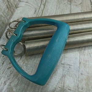 May include: A teal exercise expander with multiple silver springs. The expander has a handle with a loop on each end.