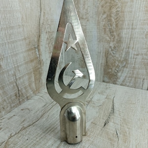 May include: A silver-colored metal finial with a cut-out design of a hammer and sickle and a star. The finial is shaped like a teardrop and has a rounded base.
