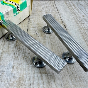 May include: Two silver-toned metal door handles with a ribbed, linear texture. Each handle features a long, rectangular bar design supported by two circular mounting bases with visible screw holes. A partial white and green cardboard box is visible.