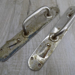 May include: Two vintage metal door handles with a silver finish and rust spots. The handles have a simple design with a curved bar and a rectangular plate.