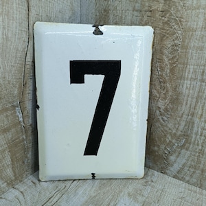 Könnte beinhalten: Weißes Email-Hausnummernschild mit schwarz lackierter Nummer 7.