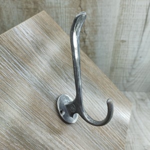 May include: A silver metal hook with a rounded base and a curved hook. The hook is mounted on a light brown wooden surface.
