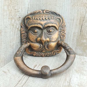 May include: A bronze lion's head door knocker with intricate details and a ring handle. The knocker is mounted on a wooden surface.