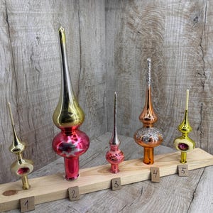 Op de afbeelding: Vijf vintage kerstboomtoppers in verschillende kleuren, waaronder goud, zilver, rood en oranje. Elke topper heeft een uniek ontwerp, met een puntige top en een decoratieve basis. Ze zijn gerangschikt op een houten display.