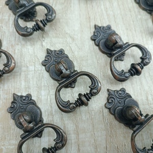 May include: A collection of ten ornate, antique-style cabinet knobs with a dark bronze finish. The knobs have a decorative, scroll-like design and a ring pull.