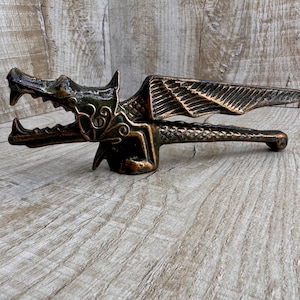 Può includere: Un schiaccianoci a forma di drago in bronzo con le ali spiegate e un design dettagliato. Lo schiaccianoci è in metallo e ha una finitura testurizzata.
