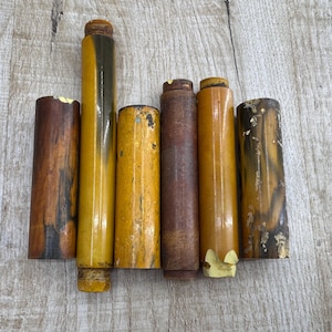 Könnte beinhalten: Eine Sammlung von sechs zylindrischen Holzobjekten in verschiedenen Braun- und Gelbtönen. Die Objekte sind vertikal angeordnet und zeigen ihre unterschiedlichen Texturen und Oberflächen. Einige haben ein glänzendes Aussehen, andere wirken gealtert.