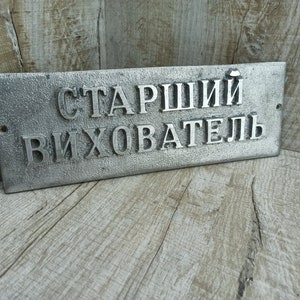Puede incluir: Un letrero de metal plateado con el texto "Старший Вихователь" en letras en relieve. El letrero est&aacute; sobre una superficie de madera.