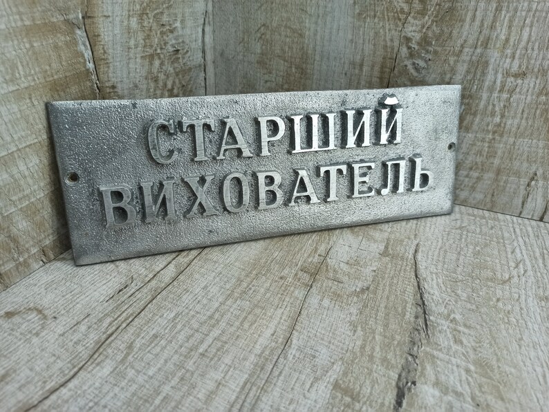 Puede incluir: Un letrero de metal plateado con el texto "Старший Вихователь" en letras en relieve. El letrero est&aacute; sobre una superficie de madera.