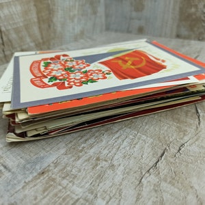 May include: A stack of vintage postcards featuring Soviet-era imagery. The postcards display vibrant colors, including red, white, and orange, with illustrations of flowers, flags, and text. The cards are stacked on a light-colored wooden surface.