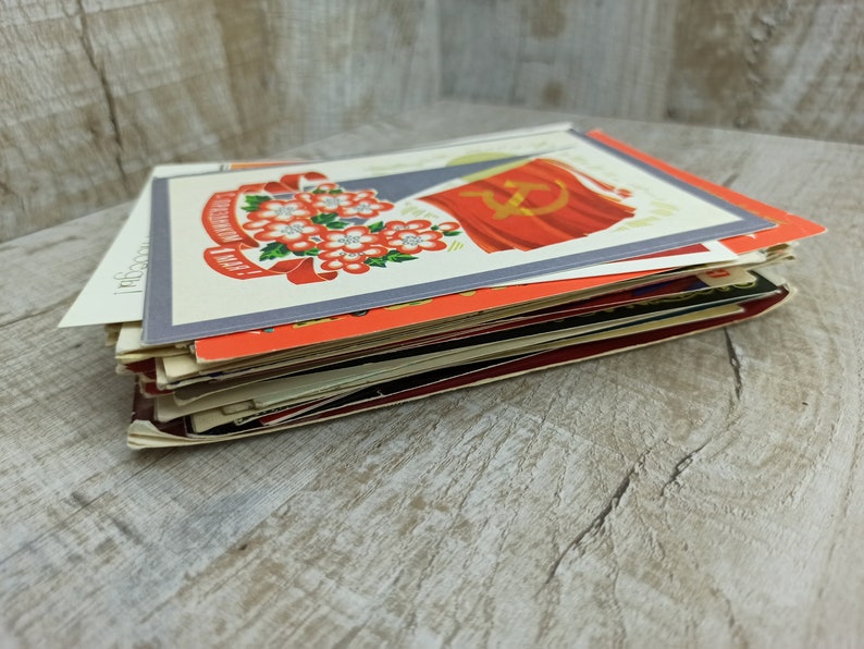 May include: A stack of vintage postcards featuring Soviet-era imagery. The postcards display vibrant colors, including red, white, and orange, with illustrations of flowers, flags, and text. The cards are stacked on a light-colored wooden surface.