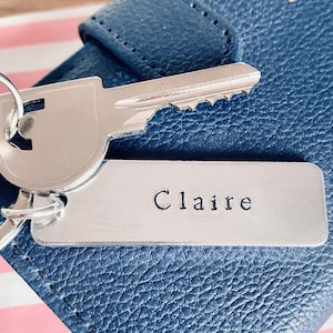 May include: A silver key chain with a silver key and a silver tag that says "Claire" on it. The keychain is attached to a blue leather wallet.