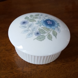 May include: White ceramic trinket box with a floral design featuring blue and purple flowers and green leaves.