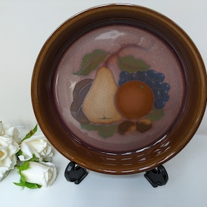 May include: A brown ceramic plate with a painted design of a pear, orange, and grapes. The plate has a raised rim and a glossy finish.