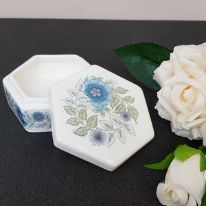 May include: A white hexagonal trinket box with a floral design in shades of blue and green. The lid is open and the inside of the box is white.