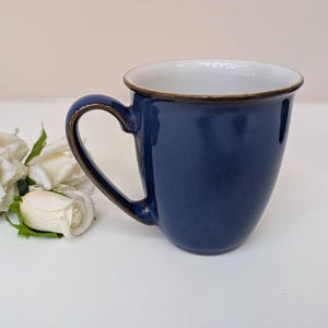 May include: A navy blue ceramic mug with a white interior and a brown rim. The mug has a curved handle and is set next to a cluster of white roses.