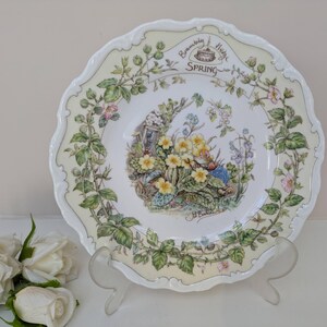 May include: A decorative plate with a scalloped edge, featuring a floral design with the words "Spring" and "Brambly Hedge". The plate has a cream border and a central image of yellow flowers and greenery. It is displayed on a clear stand.