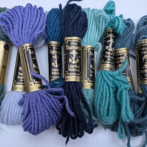 Pode incluir: Uma cole&ccedil;&atilde;o de novelos de l&atilde; Anchor Tapisserie de 10 m em v&aacute;rias tonalidades de azul e verde. Os novelos s&atilde;o envoltos em papel dourado com etiquetas pretas que apresentam um logotipo de &acirc;ncora e o texto "10m Tapisserie Laine-Wolle-Lana".
