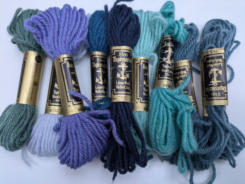 Pode incluir: Uma cole&ccedil;&atilde;o de novelos de l&atilde; Anchor Tapisserie de 10 m em v&aacute;rias tonalidades de azul e verde. Os novelos s&atilde;o envoltos em papel dourado com etiquetas pretas que apresentam um logotipo de &acirc;ncora e o texto "10m Tapisserie Laine-Wolle-Lana".