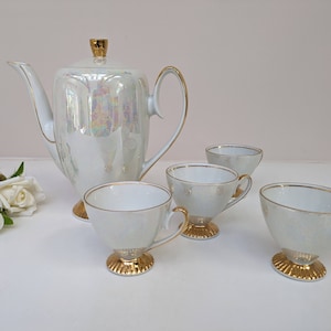 May include: A white porcelain tea set with gold accents. The set includes a teapot and four cups with saucers. The teapot has a pearlescent finish and a gold lid knob. The cups and saucers have a white finish with gold trim.