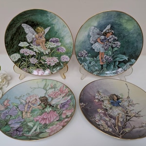 May include: Four decorative plates with fairy illustrations. Each plate has a gold rim and depicts a fairy in a garden setting with flowers. The plates are displayed on clear stands, showcasing the artwork.