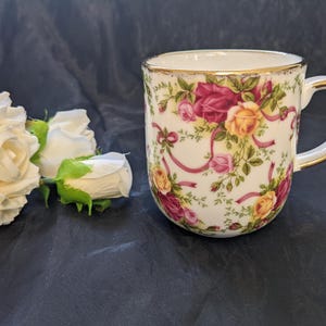 May include: A white ceramic mug featuring a floral pattern of red, pink, and yellow roses with green leaves and pink ribbons. The mug has a gold-trimmed rim and a gold-trimmed handle. White artificial roses are positioned to the left of the mug.