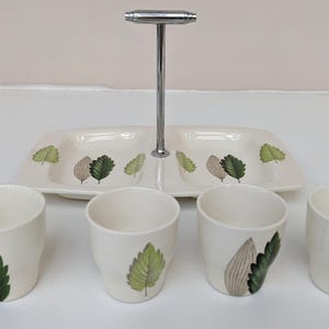 May include: A white ceramic serving tray with a metal stand and four small cups. The tray is divided into two sections and features a leaf design. The cups also have a leaf design.