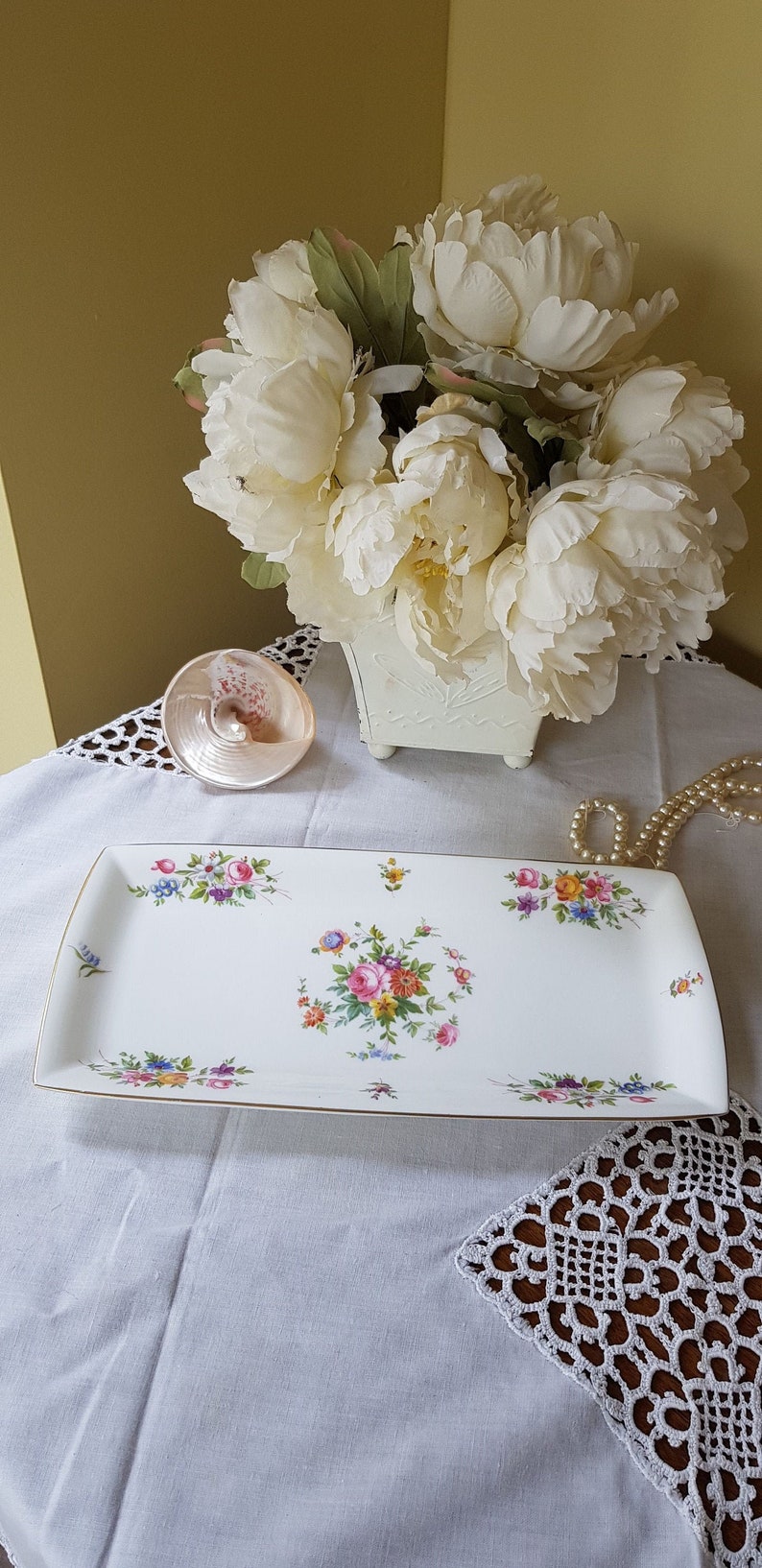 May include: A white rectangular serving tray with a gold trim and a floral design. The tray is on a white tablecloth with a crocheted lace doily.