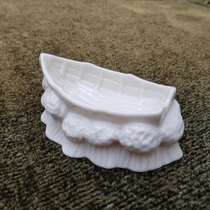 May include: A white ceramic boat-shaped dish with a textured, leafy design around the base. The dish is designed to look like a boat resting on a bed of leaves.