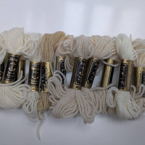 Pode incluir: Oito novelos de l&atilde; Anchor Tapestry em v&aacute;rios tons de branco e creme. A l&atilde; est&aacute; enrolada em carret&eacute;is de cart&atilde;o com etiquetas douradas que dizem "Anchor Tapestry Laine a Broder" e apresentam um logotipo de &acirc;ncora.