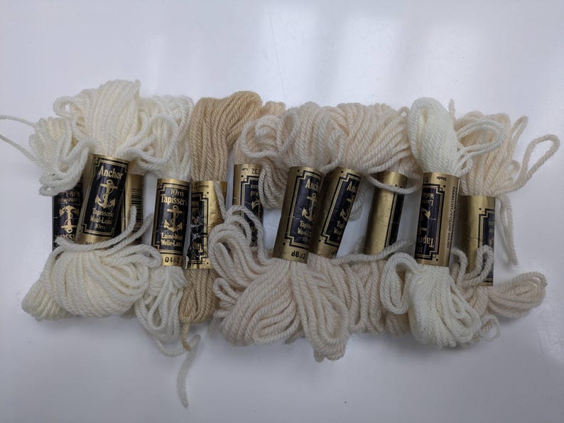Pode incluir: Oito novelos de l&atilde; Anchor Tapestry em v&aacute;rios tons de branco e creme. A l&atilde; est&aacute; enrolada em carret&eacute;is de cart&atilde;o com etiquetas douradas que dizem "Anchor Tapestry Laine a Broder" e apresentam um logotipo de &acirc;ncora.