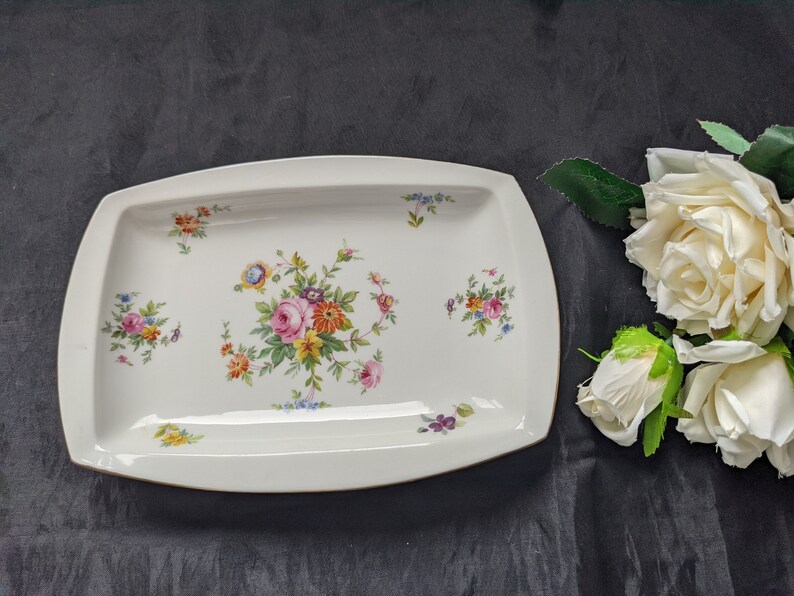 May include: A white rectangular serving platter with a floral design. The platter has a raised rim and a gold trim. The flowers are pink, yellow, and blue.