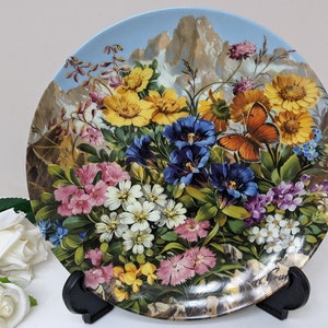 May include: A decorative plate with a colourful floral design, including yellow, blue, pink, and white flowers, and an orange butterfly. The background shows a mountain range under a light blue sky. The plate is on a black stand.