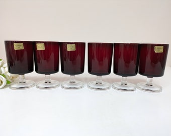 May include: Set of six clear glass stemmed wine glasses with a dark red base. The glasses have a gold label with the word "Lumineux" printed on them.